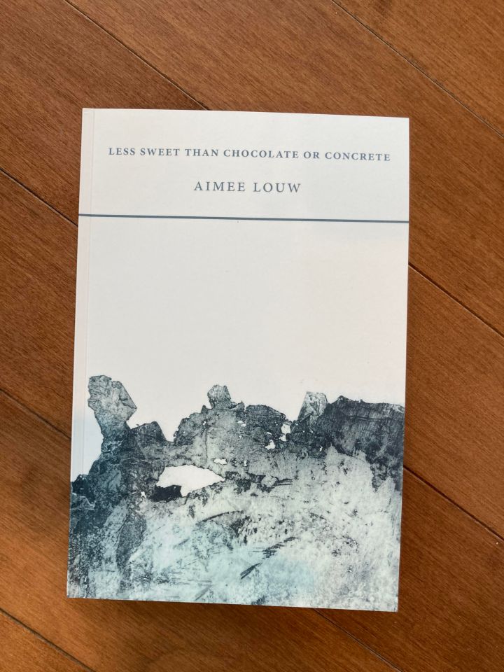 Image description: Image is the cover of a white book with the title, Less Sweet than Chocolate or Concrete, at the top, and an image resembling a tideline with asymmetrical spaces of dark-blue-grey and spaces of white covering the bottom half of the page. The book is placed on warm, honey brown wood panels.